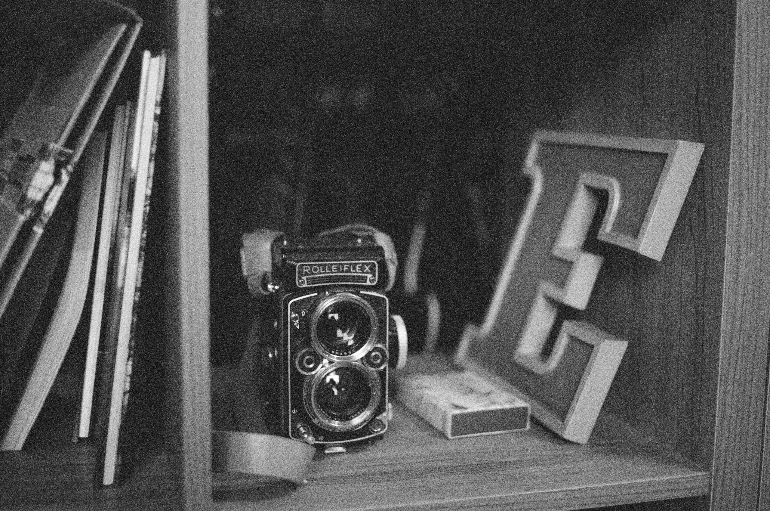 A bookshelf with books, a Rolleiflex camera, and a metal letter E